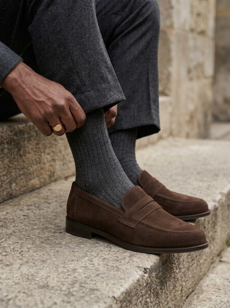 Hombre sentado en un escalón subiéndose el calcetín con los mocasines Estribo de ante color tabaco