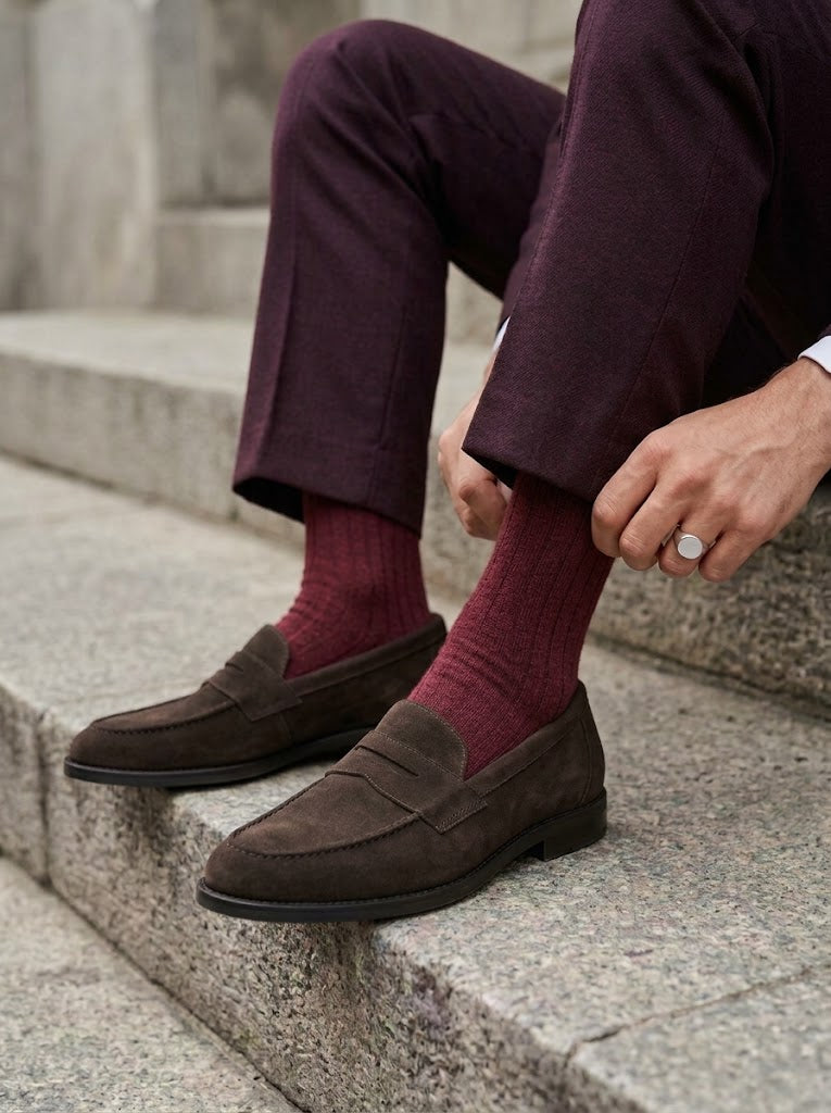 Hombre sentado en un escalón subiéndose el calcetín con los mocasines Estribo de ante color chocolate