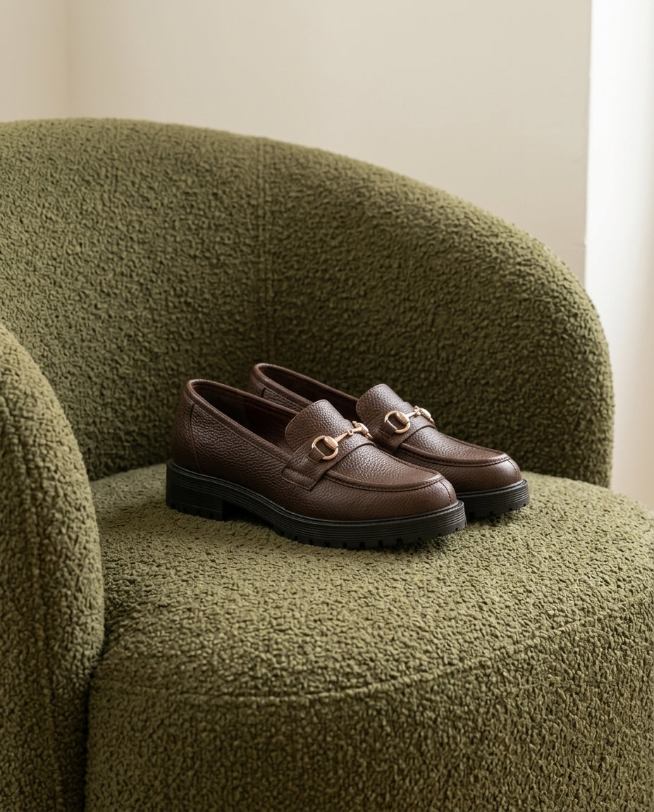 Mocasín Traviesa chocolate, sobre el asiento de una butaca de bouclé verde oliva