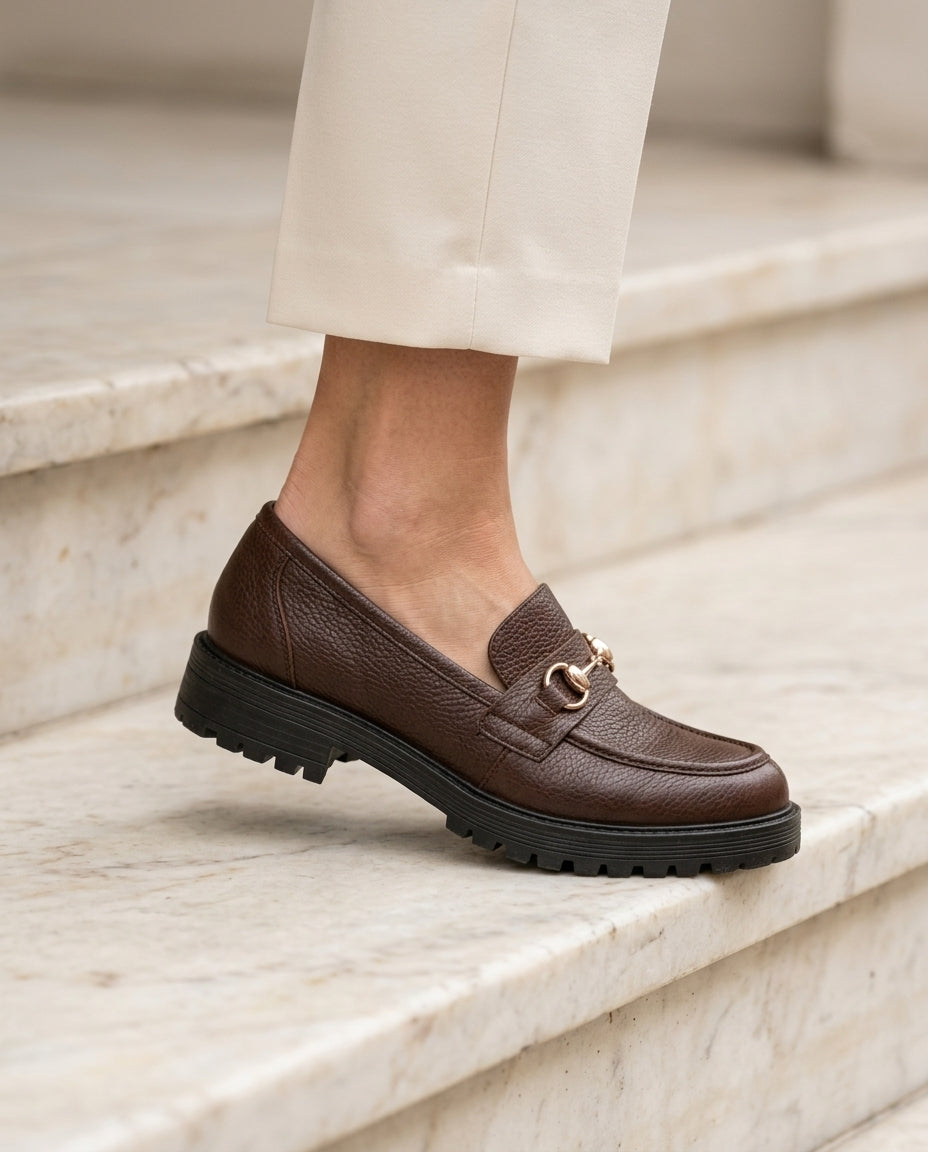 Mocasín Traviesa chocolate, pisando un escalón de mármol con pantalón crema