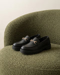 Mocasín Traviesa negro, sobre el asiento de una butaca de bouclé verde oliva