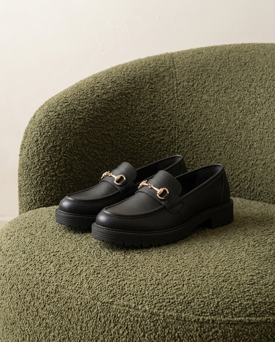 Mocasín Traviesa negro, sobre el asiento de una butaca de bouclé verde oliva